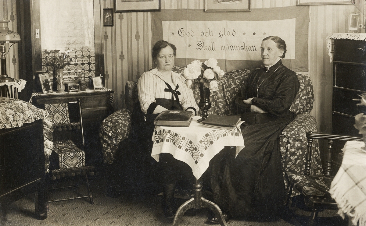 Två kvinnor sitter i en soffa i ett vardagsrum. Framför sig har de ett pelarbord med några fotoalbum.