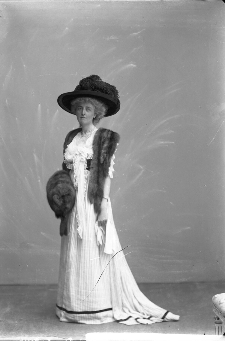 Porträtt av en kvinna i ljus lång klänning med boa runt halsen, muff och vidbrättad hatt.