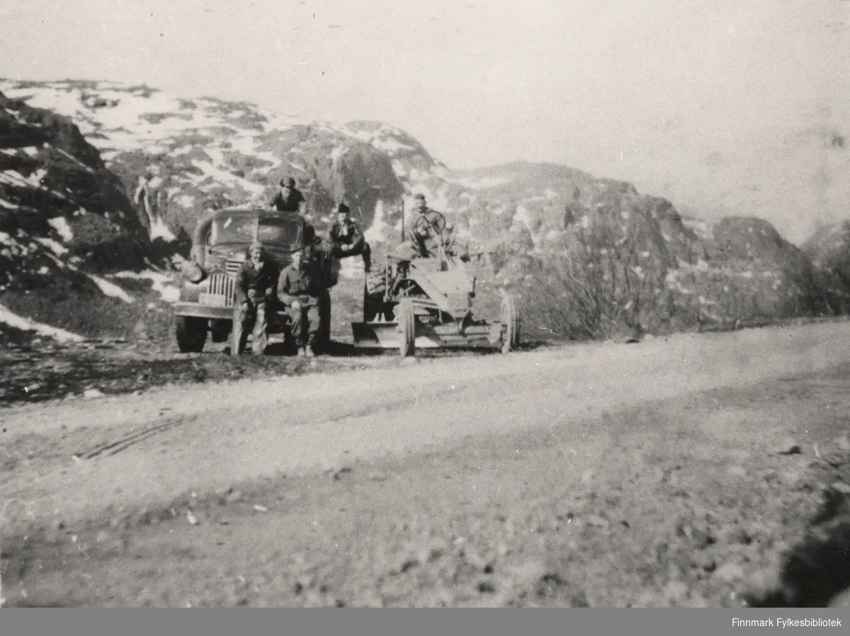 Lastebil og veiskrape. Sjåfør Karl Labbahå sitter til høyre på støtfangeren til lastebilen. De fire andre mennene er ukjente. Bildet er tatt på Brannseltta i Bugøyfjord, Sør-Varanger.