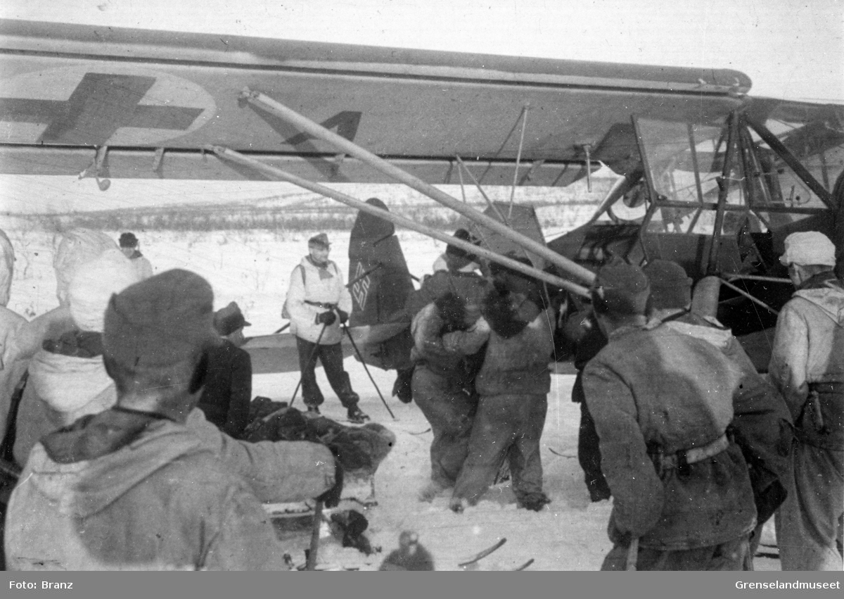 Transport av sårede soldater fra en skitropp. En gruppe soldater i vinteruniform med ski og staver ser på at en båre blir plassert inn i  flyet som er merket med både hakekors og Røde Kors. Flyet er et Fieseler Storch ambulansefly.