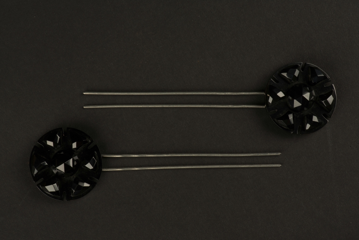 Två hårnålar med rund dekoration i svart.
Nålarna består av parallella långa hårnålar samt en dekoration i form av en mönsterslipad svart platta. Denna är troligen tillverkad i plast, eventuellt cellulosaacetat.