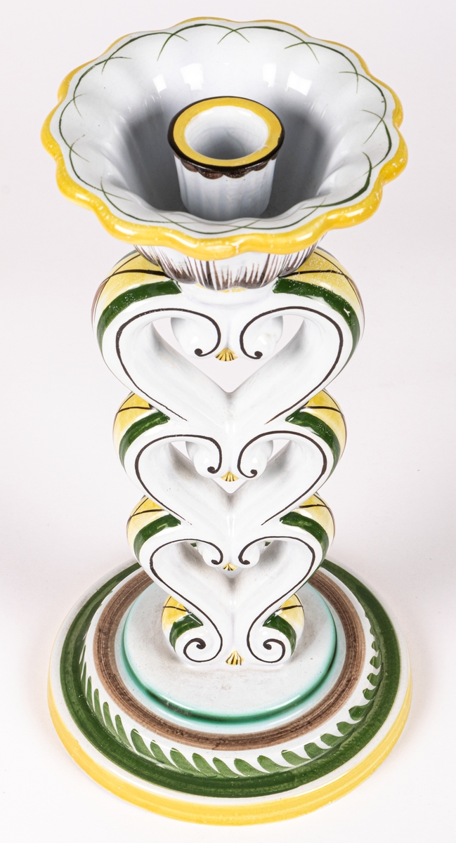 Ljusstake modell H från Gefle Porslinsfabrik, formgiven av konstnär Arthur Percy 1926 för fabriken. Rund fotplatta som bär upp tre på varandra stående stiliserade hjärtan, överst en ljushållare i form av en öppen blomma. Ljusstake H har tillverkats med olika glasyrer. Just denna är handmålad i dekor 64 av chefsdekoratör Eugen Trost, som signerat med T. på undersidan.