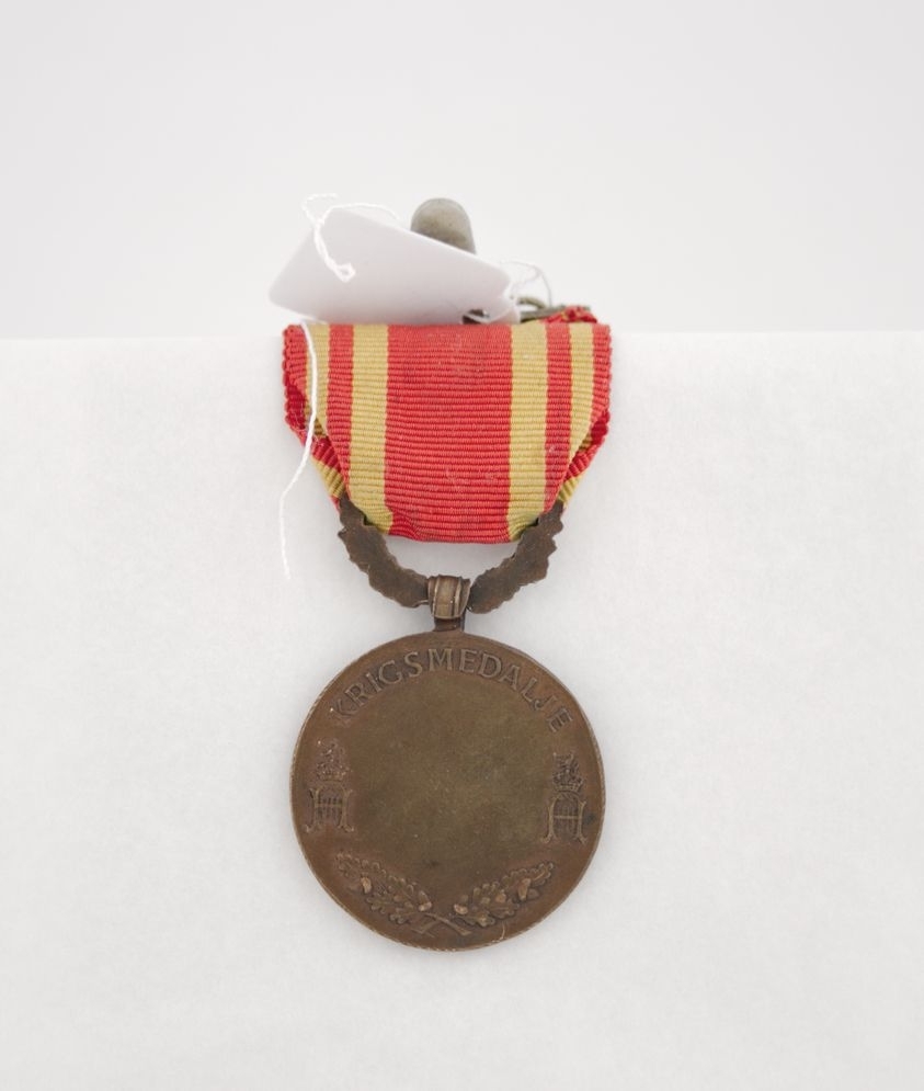 Medalje i bronse med portrett av Haakon VII. Dekorert med eikekrans, korslagte eikegreiner og kongens monogram. Gult og rødt bånd.