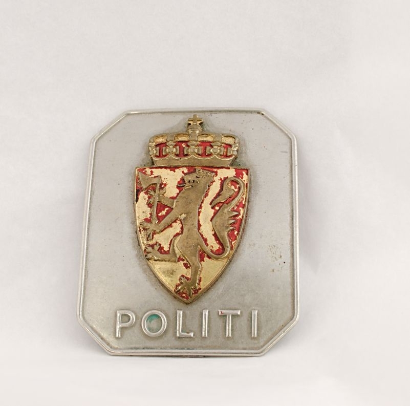 Åttekantet polititegn i nysølv med riksvåpenet av 1937 og teksten "POLITI". Den røde emaljen i riksvåpenet er slitt.

