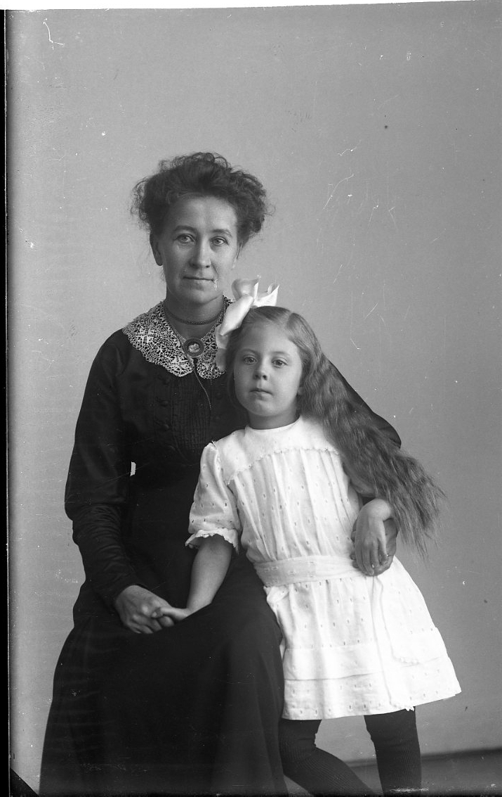 En kvinna sitter på en stol med en flicka stående vid sin sida. Troligen mor och dotter. Kvinnan är fru Kristiansson i Gränna.