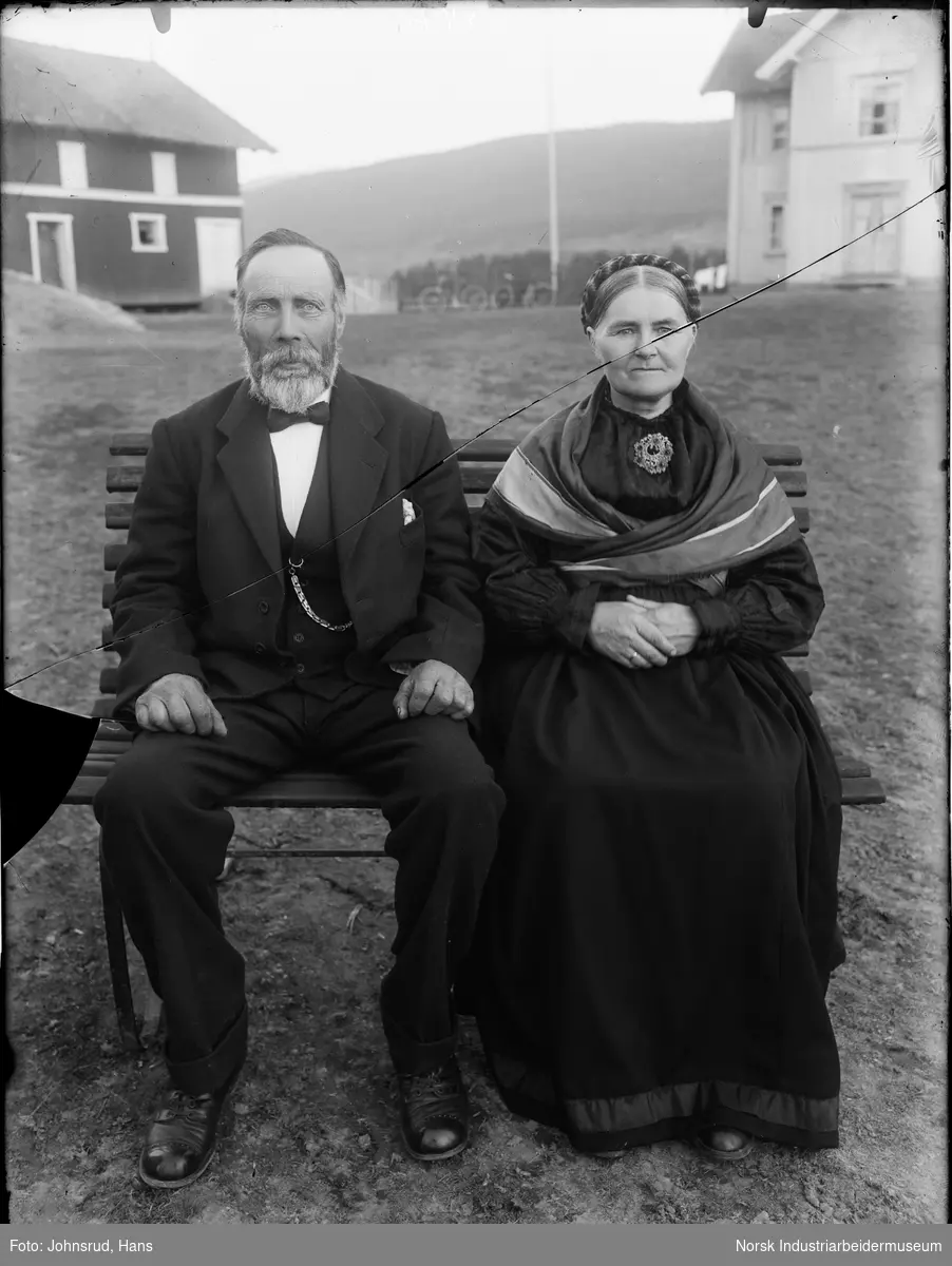 Portrett av mann og kvinne som sitter på en benk i gårdstunet. I bakgrunnen til venstre er det en låve, i midten en flaggstang og to sykler, og til høyre et bolighus.