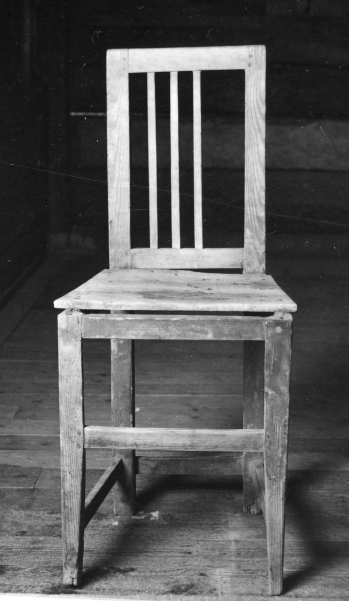 Omålad stol med raka 4-sidiga ben, tvärslåar, rektangulär rygg med 4 vertikala spjälor varav en saknas.