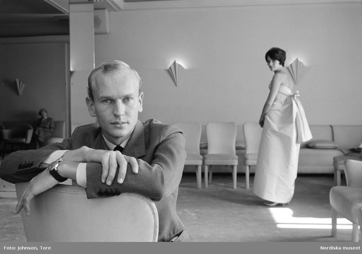 Porträtt av en man i interiör med en stående kvinna i bakgrunden. Palle Wikberg i salongen på NK:s Franska damskrädderi, hösten 1960. Klänningen i rosa är en originalmodell från Cristóbal Balenciaga.