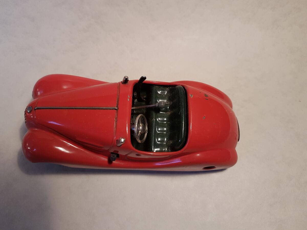 En röd leksaksbil för två personer. Den kommer från leksaksföretaget Schuco. Modell Examico 4001. Tillverkad i: US-Zone Germany. Tidigare uppdragbar, nyckel saknas. Vindruta saknas.