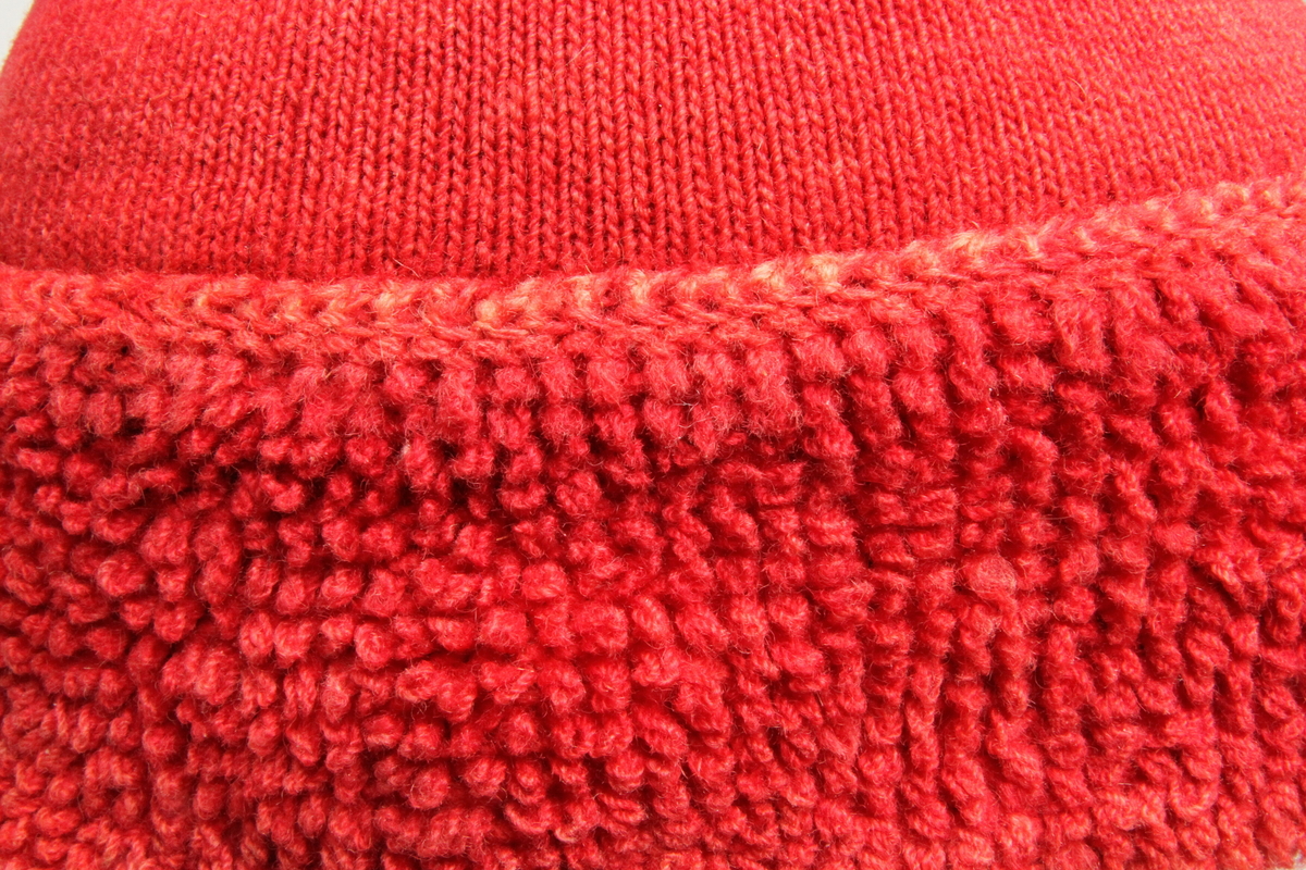 Rød strikket topplue. Lua er tett strikket med små pinner og forholdsvis tynt, hardt spunnet garn. Med flosset 8,5 cm bred kant nederst, ant. strikket i løkkestrikk. Liten dusk på toppen. Sammensydd i spissen. Foret med naturhvit strikket for. 