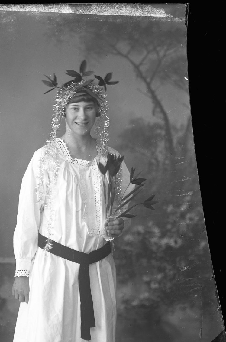 Porträtt av en ung kvinna i ljust lång linne med blommor och glitter i håret och blommor i handen. En lucia?