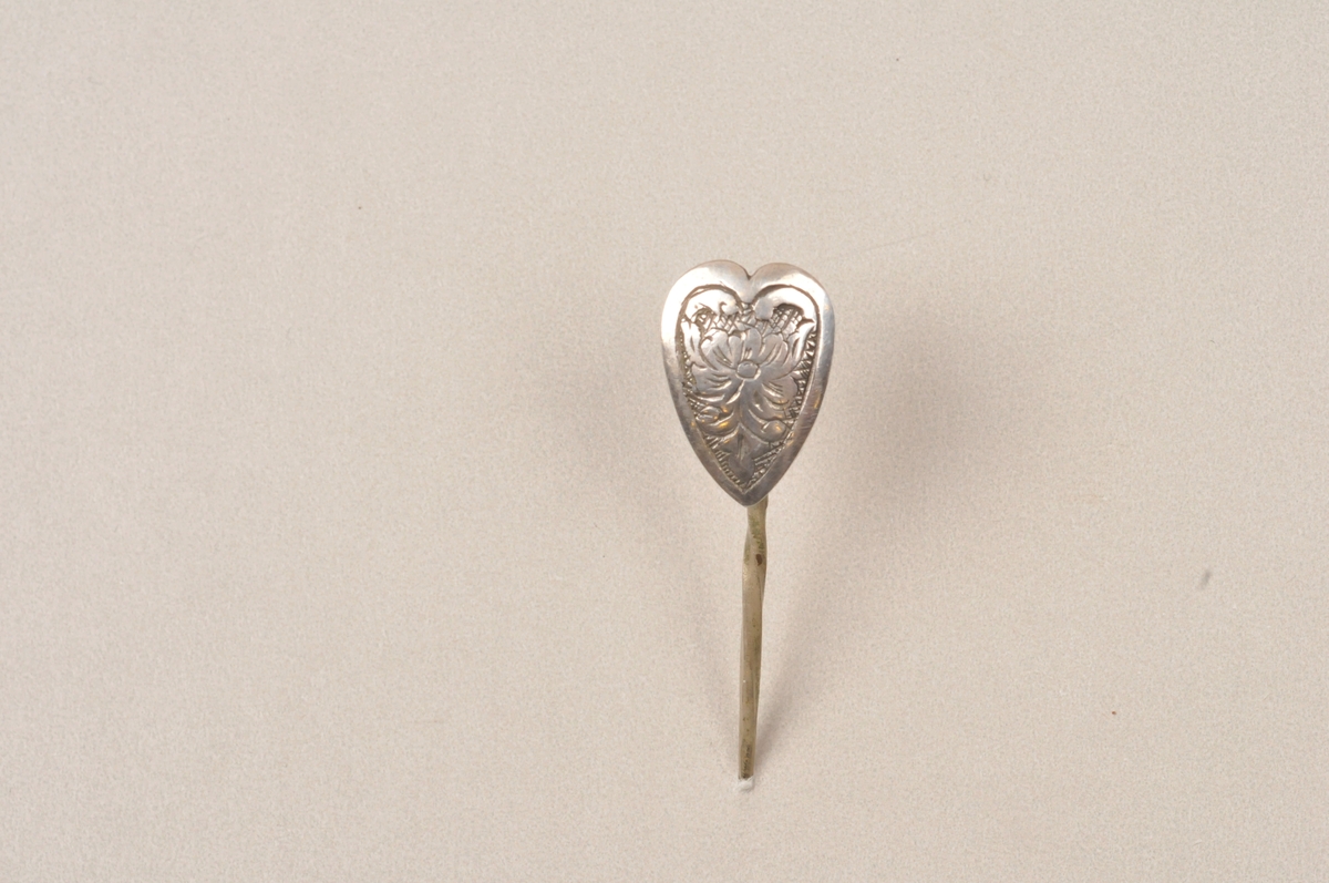 Hjerteforma plate med gravera dekor og pålodda nål på baksida.