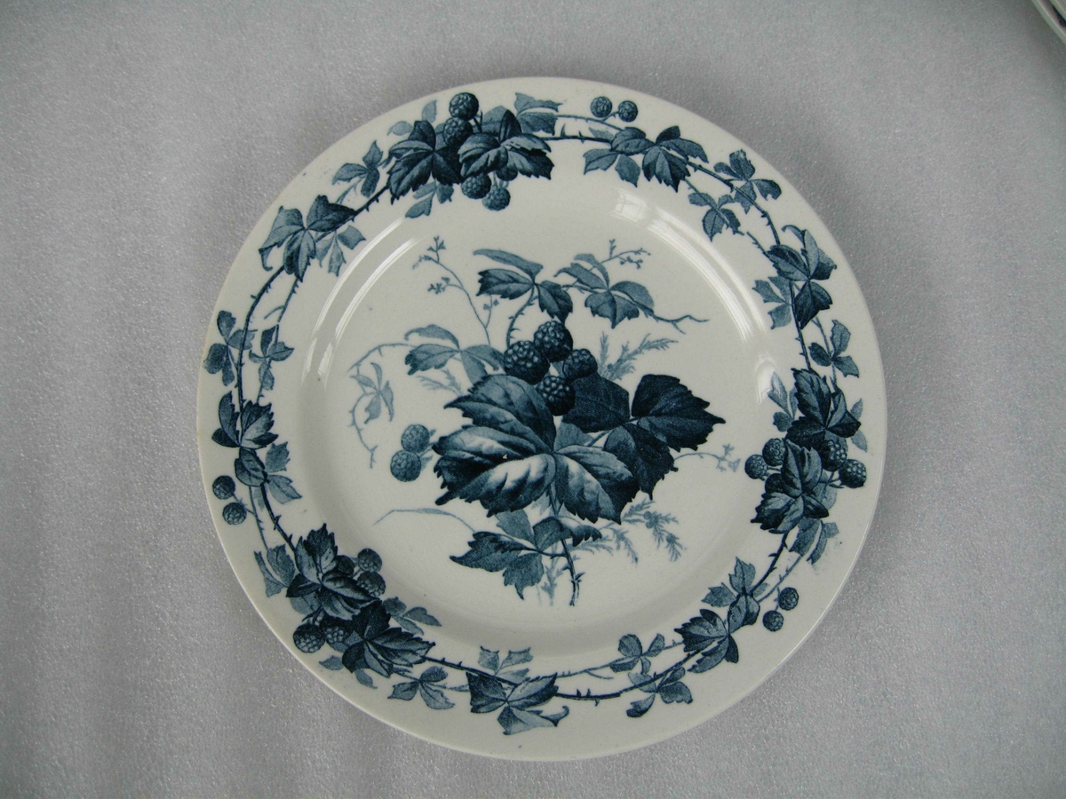 Asjett av hvit keramikk med blå dekor av bjørnebærmotiv. En ranke med blad og bær langs kanten og en kvist med blad og bær i bunnen.