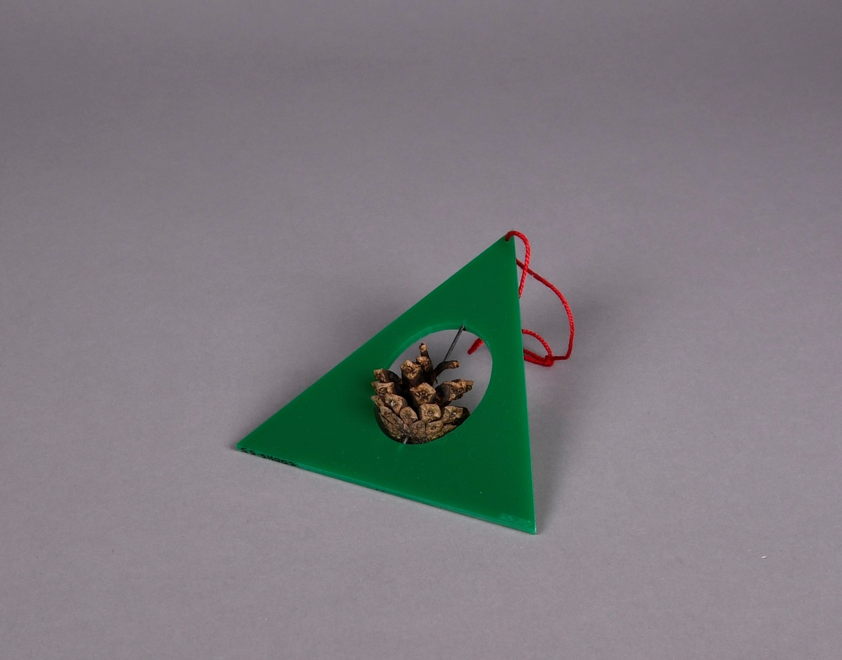 Juletrefigur av farget akryl og furukongle. Juletre formet som grønnfarget trekant med ovalt hull i midten, i hullet er det en stav av metall som går gjennom en furukongle.
