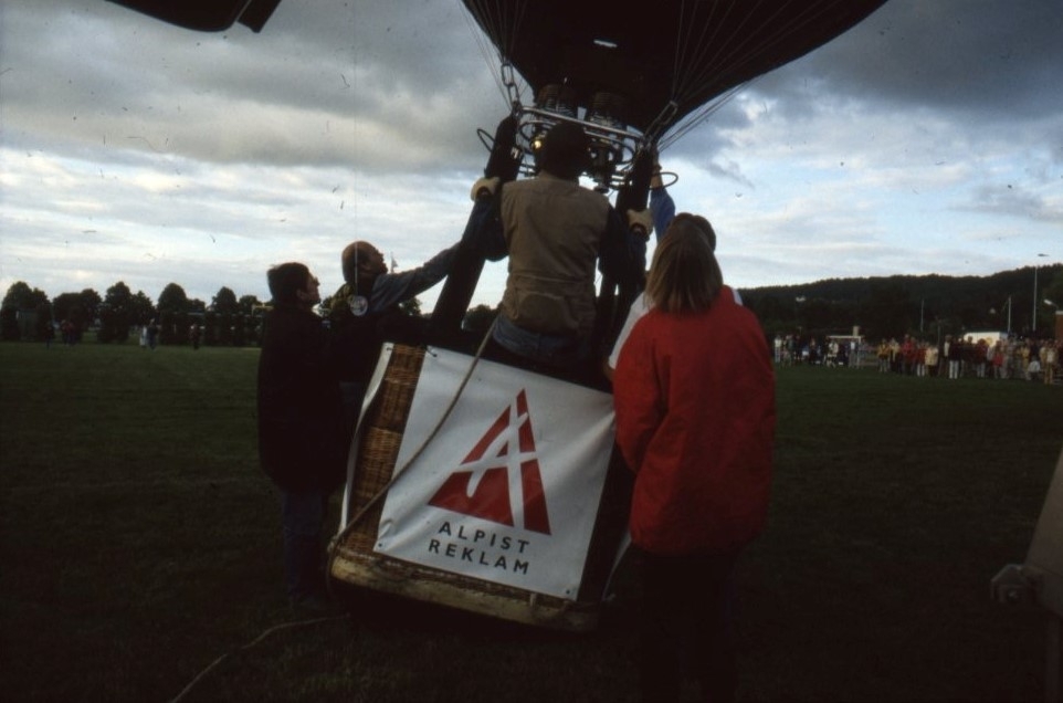 Fyra personer står och håller i en ballongkorg som håller på att lyfta. I korgen står en man och håller i sig. På ballongkorgen står det Alpist reklam.