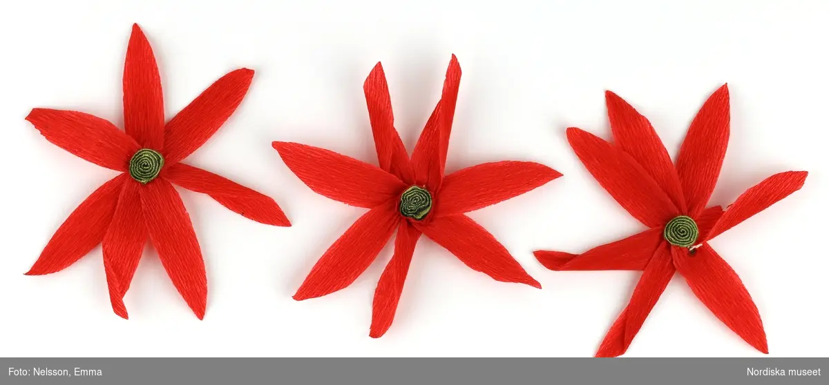 a-l) Tolv stycken hemtillverkade julgransprydnader av rött kräpp-papper i form av blommor/julstjärnor, med upphängningsanordning av knappnålar. 

Lena Kättström Höök 2019-03-21