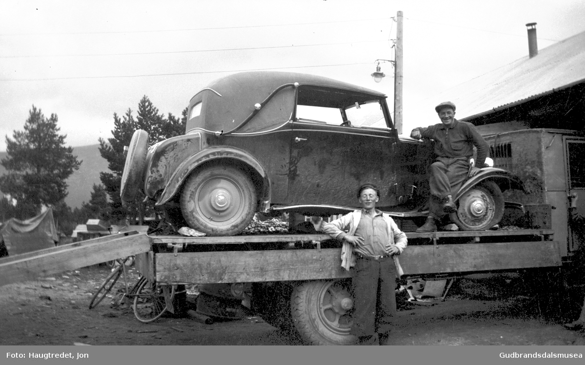 Ein personbil har vorte transportert til bilverkstaden i Bismo på ein lastebil. 
Arthur Aurmo (f. 1906) står framfor og Mathias Moastuen (f. 1912) sit på lasteplanet
