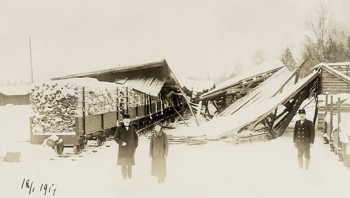 Foto av ett sågverk där det verkar ha ha hänt en olycka. 18/1 1917.
Två män och en kvinna inspekterar förödelsen.