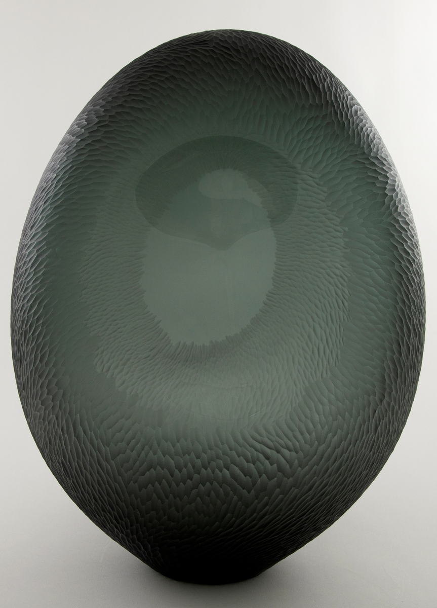 Noe flattrykket ovalformet skulptur i gråfarget klart glass. Deler av overflaten er behandlet med sliping, sandblåsing og polering, slik at det oppstår en matt bølgende struktur over store deler av vasen. Midten av de konkave langsidene er ubehandlet, noe som muliggjør at betrakteren kan studere dekoren fra både ut- og innsiden. Sirkulær åpning i vasens bunn.
