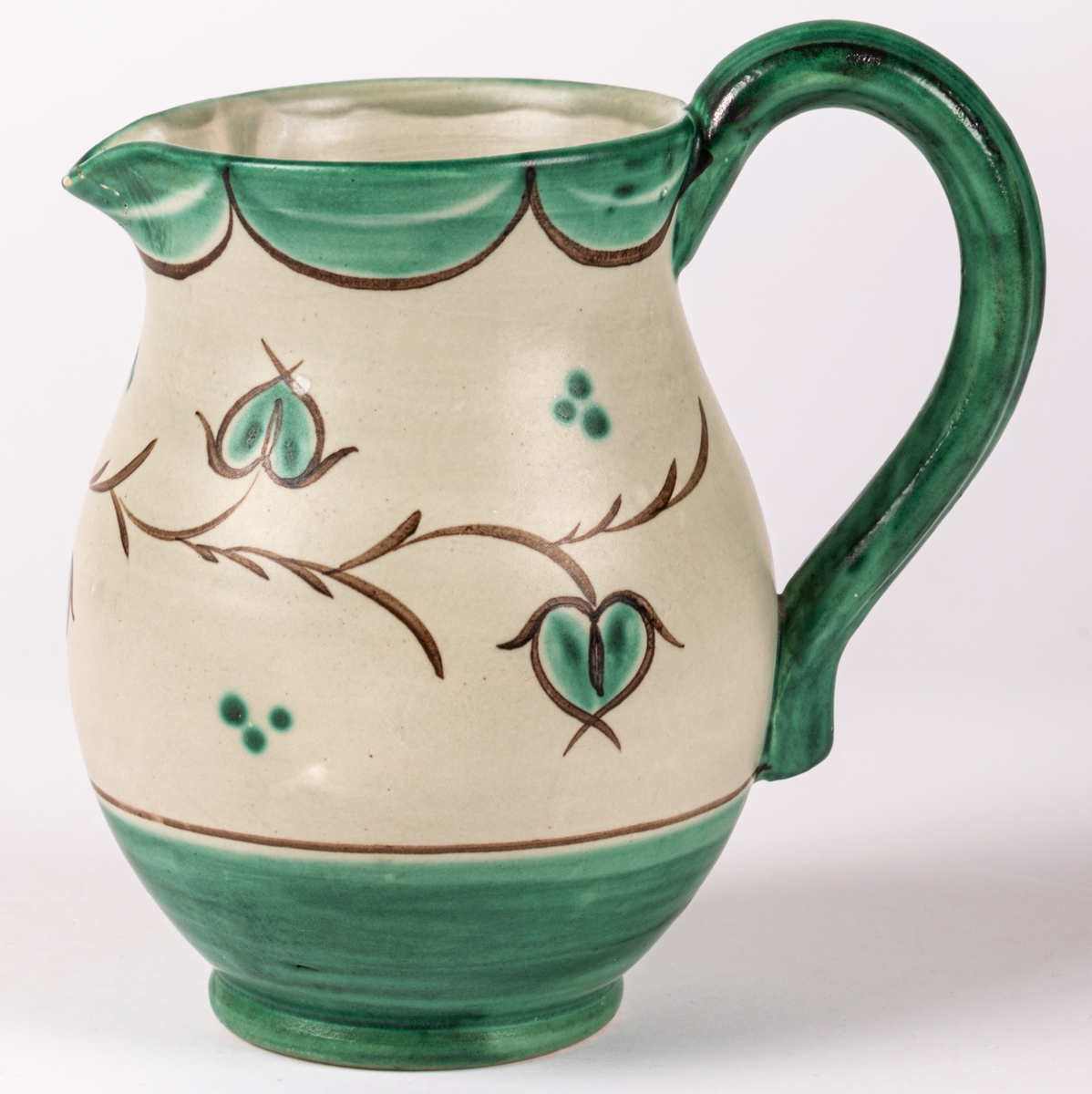 Tillbringare med hänkel i lergods från Bo Fajans, rymmer 3,5 dl. Formen skapad 1925 av konstnär Eva Jancke-Björk. Denna dekor/glasyr VA - grågrön matt glasyr med grön och brun dekor - producerades mellan 1935-43. Pipen limmad.