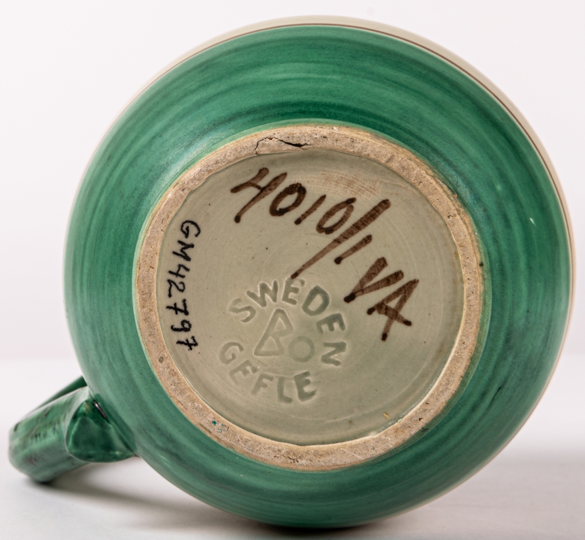 Tillbringare med hänkel i lergods från Bo Fajans, rymmer 3,5 dl. Formen skapad 1925 av konstnär Eva Jancke-Björk. Denna dekor/glasyr VA - grågrön matt glasyr med grön och brun dekor - producerades mellan 1935-43. Pipen limmad.