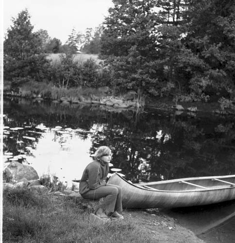 En kvinna sitter vid en å. Bredvid henne ligger en kanot uppdragen på land.