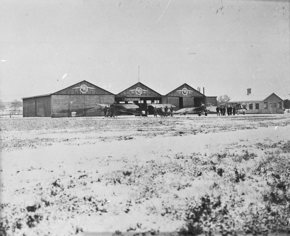 Exteriör av Thulins flygskola i Ljungbyhed med hangar och verkstäder, vintertid, omkring 1918. Från vänster står flygplan Thulin A nr 7, Thulin A nr 4, Thulin D, Thulin B samt två flygplan SW 15.