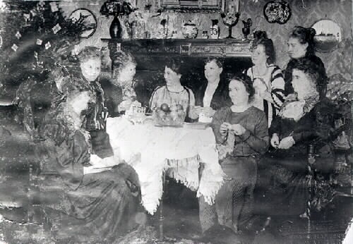 Nio handarbetande damer runt ett bord vid julgranen. Kanske är det ett symöte eller något julfirande hos Alida (eller hos Mathilda Ranch?) Beställare: Alida Svensson.