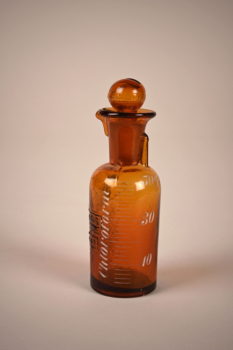 Flaske i brunt glass med propp. Målestreker er malt på i hvitt. 10, 30 og 50 gram er merket med tall på flasken.