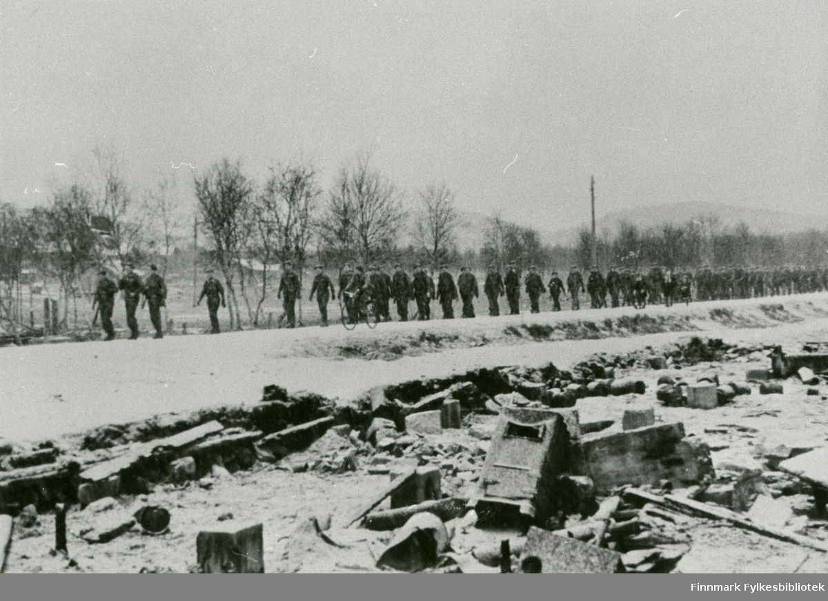 Norske soldater på norsk jord, Bergkompaniet marsjerer mot Bjørnevatn med norsk flagg i hånden til de første i marsjen, 1944. Vi ser ruiner i forgrunnen.
