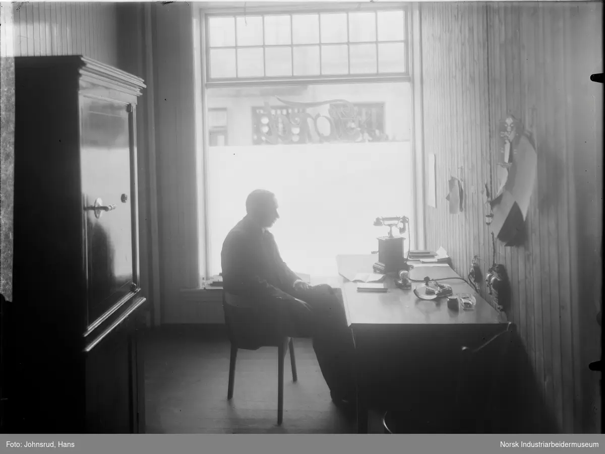 Mann sittende ved skrivepult foran vindu. Ordet "Ford" er synlig speilvendt i vinduet. Det står en telefon på bordet.