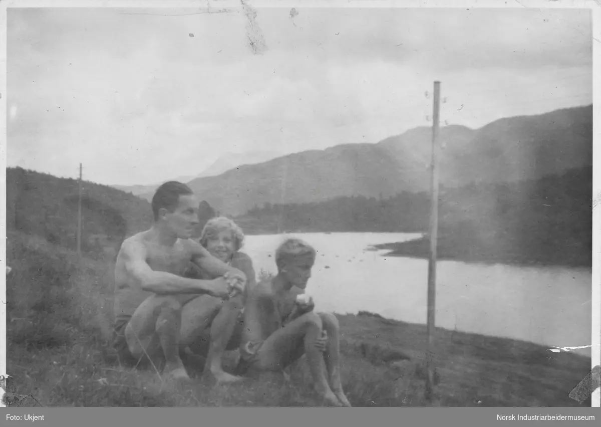 Far med to barn sittende på bakken ved Hyldalen seter. Hyldalsvannet i bakgrunnen.