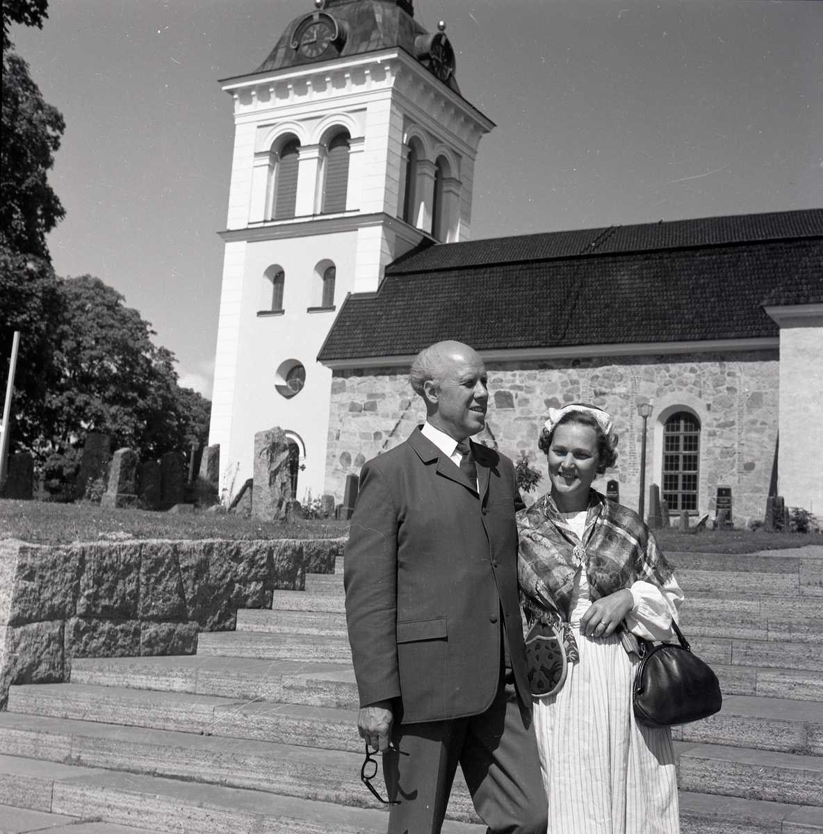 En man i kostym står tillsammans med en kvinna i hembygdsdräkt på en trappa nedanför en kyrka.