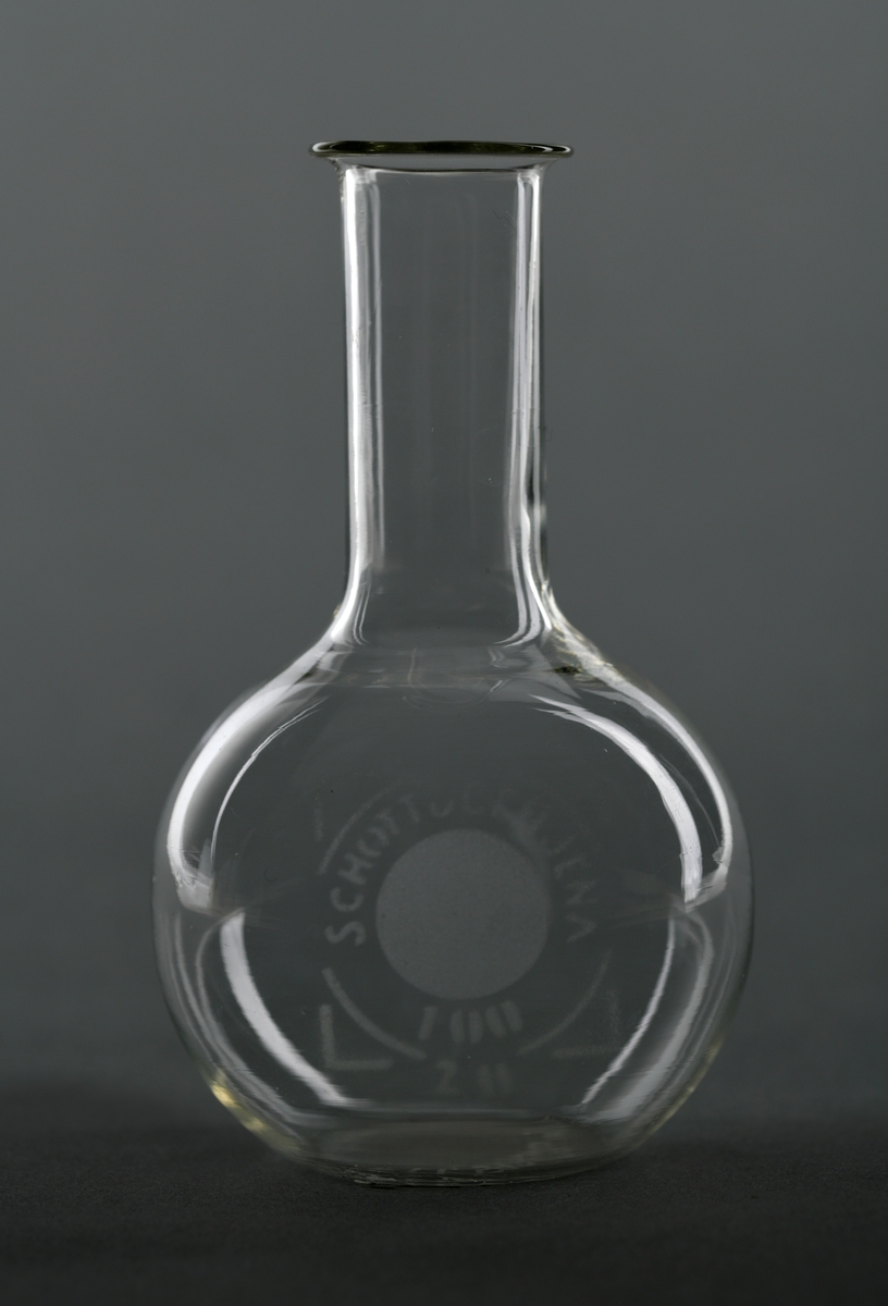 En målekolbe av glass, trolig borosilikatglass. Glasskolben har rund form med lang og relativt tynn hals. Ofte blir slike kolber brukt til å lage løsninger med et visst volum og konsentrasjoner.
