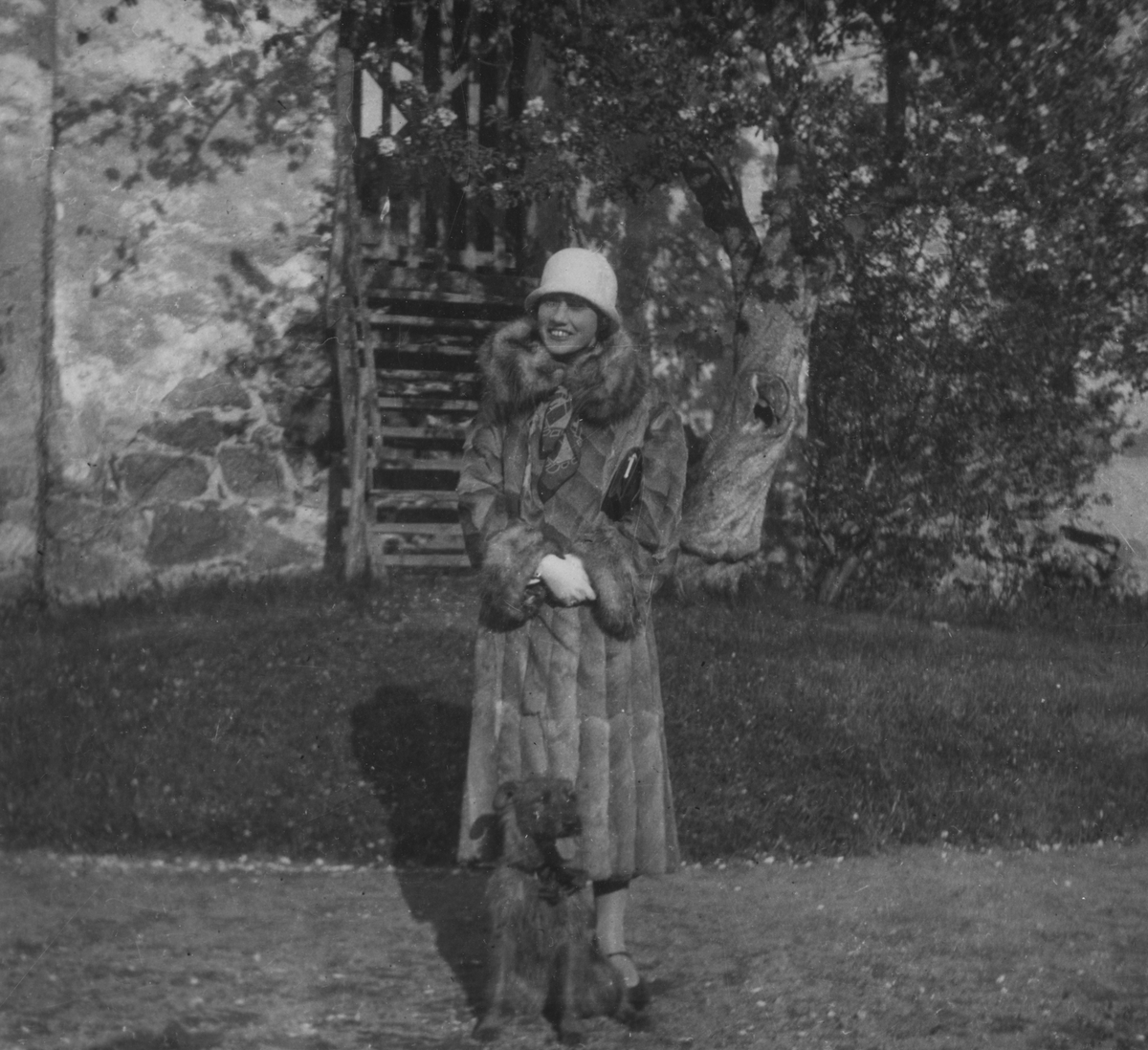 Porträttfoto av Anna Linderstam iklädd rock och hatt med en hund framför en byggnad i Stjärnorp, 1925.

Text vid foto: "Med Fiat 501 på strövtåg i vida världen. 1925. Flickan vid Stjernorp. BK."