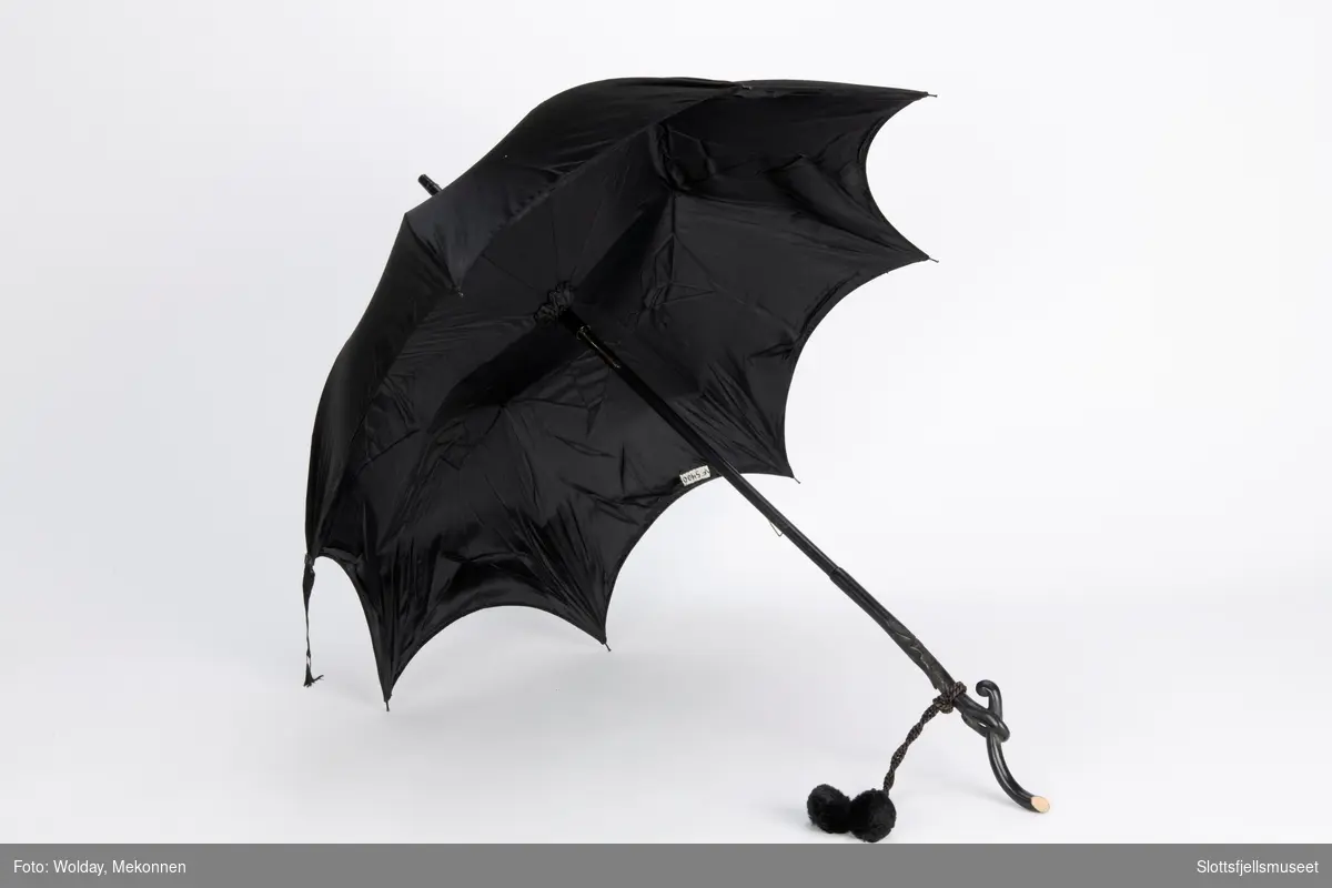 Paraply med 8 spiler av metall og svart treskaft med utskjæringer. Ved håndtaket er festet en tvunnet snor med  pomponger.  Paraply er trukket og fôret med svart stoff.