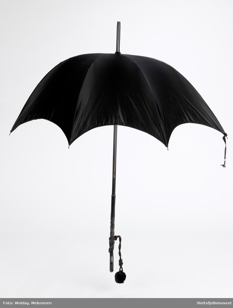 Paraply med 8 spiler av metall og svart treskaft med utskjæringer. Ved håndtaket er festet en tvunnet snor med  pomponger.  Paraply er trukket og fôret med svart stoff.