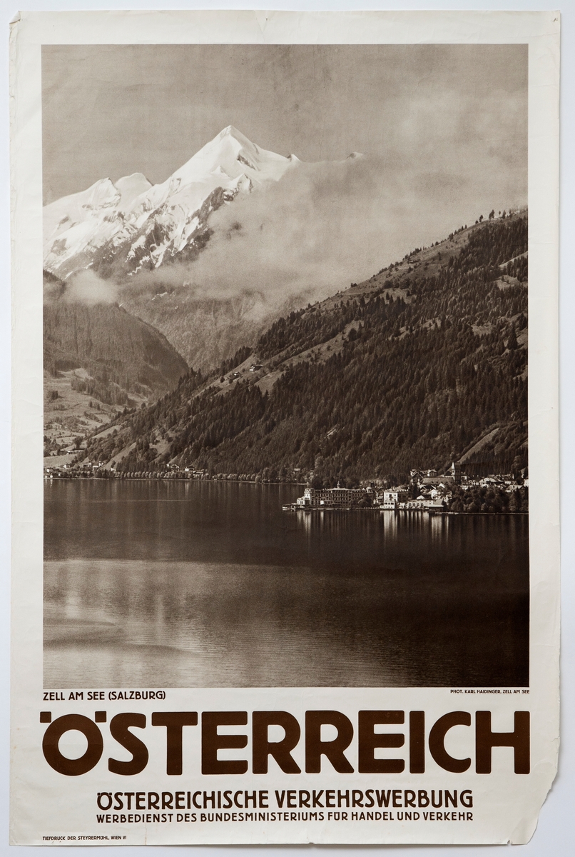 Gjengivelse av et fotografi med sjø i forgrunnen og bebyggelse til høyre under bratte fjellsider. I bakgrunnen er det fjell med snø, delvis skjult av tåke.