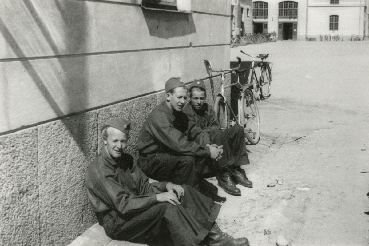 Tre soldater sitter vid en kasernbyggnad i Umeå, 1940.

Troligen på området för K 4, Norrlands dragonregemente.

Bildtext vid foto: "Umeå den 18.6 1940 Brrr!"

Foto skannat ur album: 'Minnen från Min kommistid 1940-1944'