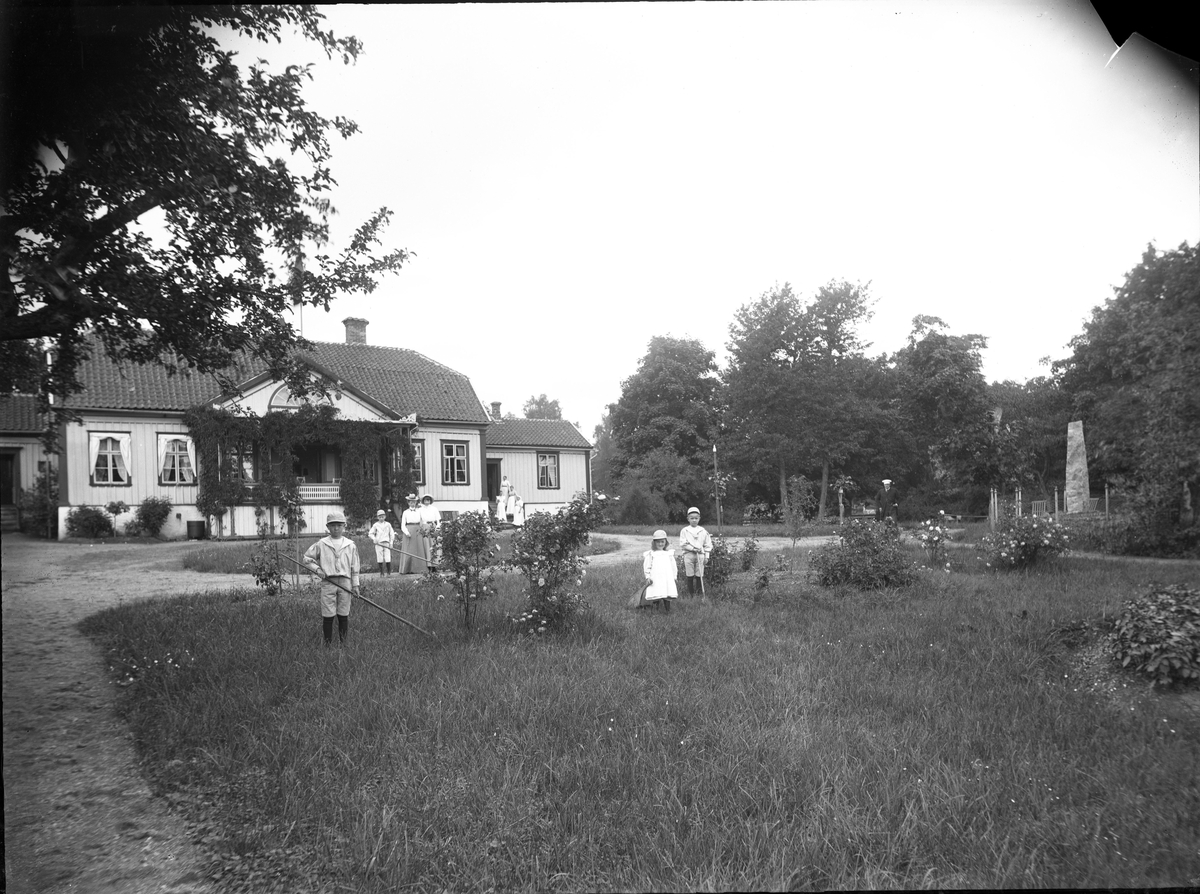 Barn och kvinnor på en gårdsplan till en herrgårdsliknande byggnad. Till höger en hög minnessten/bautasten.
