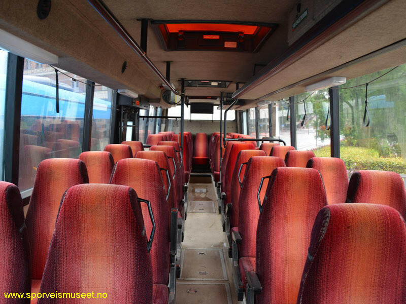 Grønn buss med to inngangspartier, en fremme og en i midten. Bussens interiør består av gulv og tak i en beige farge og røde og oransje seter. 