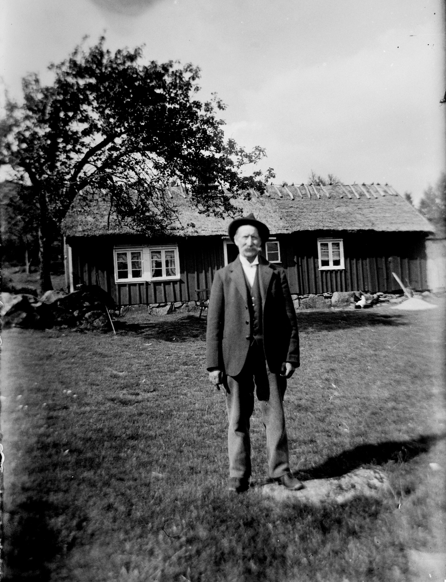 Gamle Frans Hallengren står i hatt och kostym på gräsbacken framför stugan på Långarekullen.