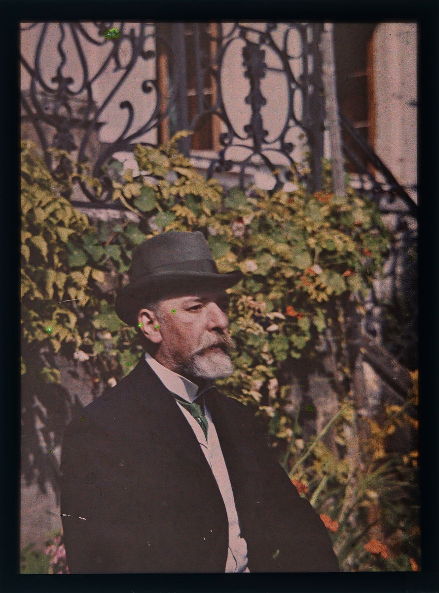 Portrett av en mann iført dress og hatt foran en husvegg med planter.