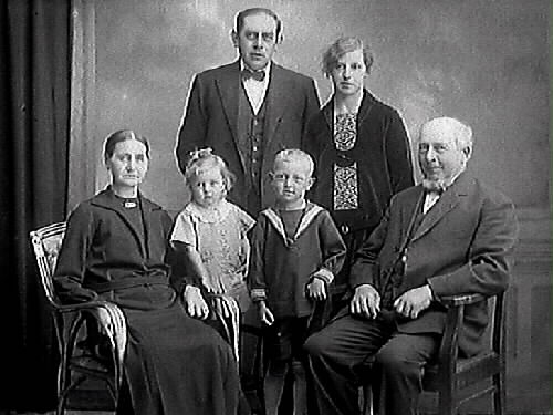 Familjebild. Familj från Träslöv. Kan möjligen vara tre generationer med det äldsta paret sittande, ett medelålders par stående och deras två barn.