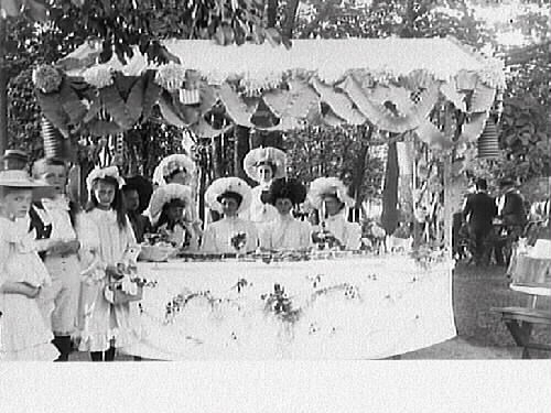 Sju flickor i ymniga hattar i ett marknadsstånd, rikt utsmyckat med pappersdekorationer. Eventuellt en basar eller firande av Barnens dag.