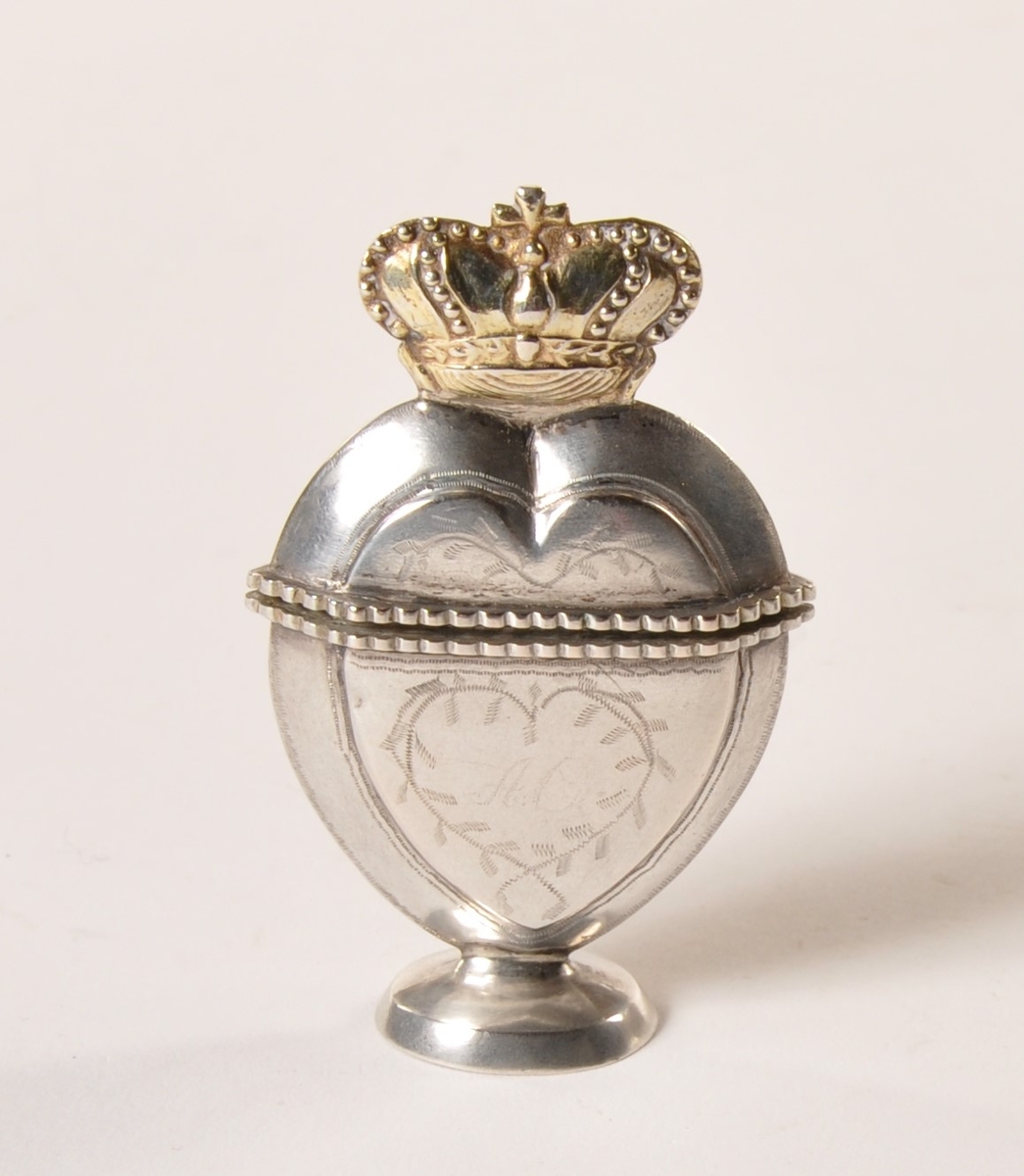 Hjerteformet luktevannshus i sølv  med henglset lokk med  gullfarget krone. Stående på sokkel. Ingravert bladmønster.