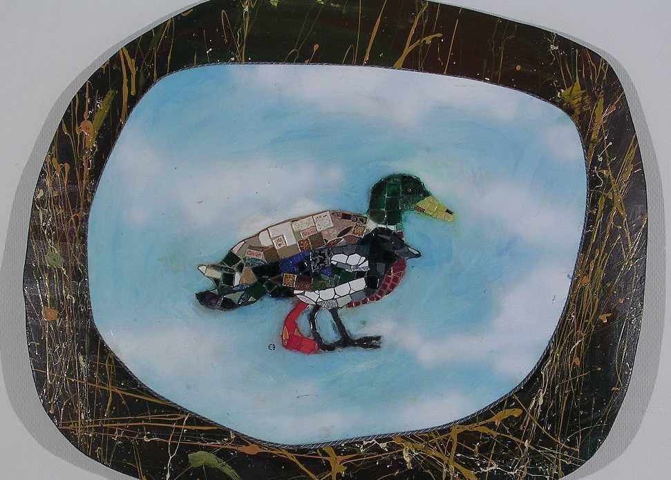 På en ojämn rund pannå med brun bakgrund med gula, grön och orange färgstänk  sitter en tjockare pannå målad i himmelsblå färger. Ovanpå denna två fåglar i mosaik. Runt den tjockare pannån går ett rep.