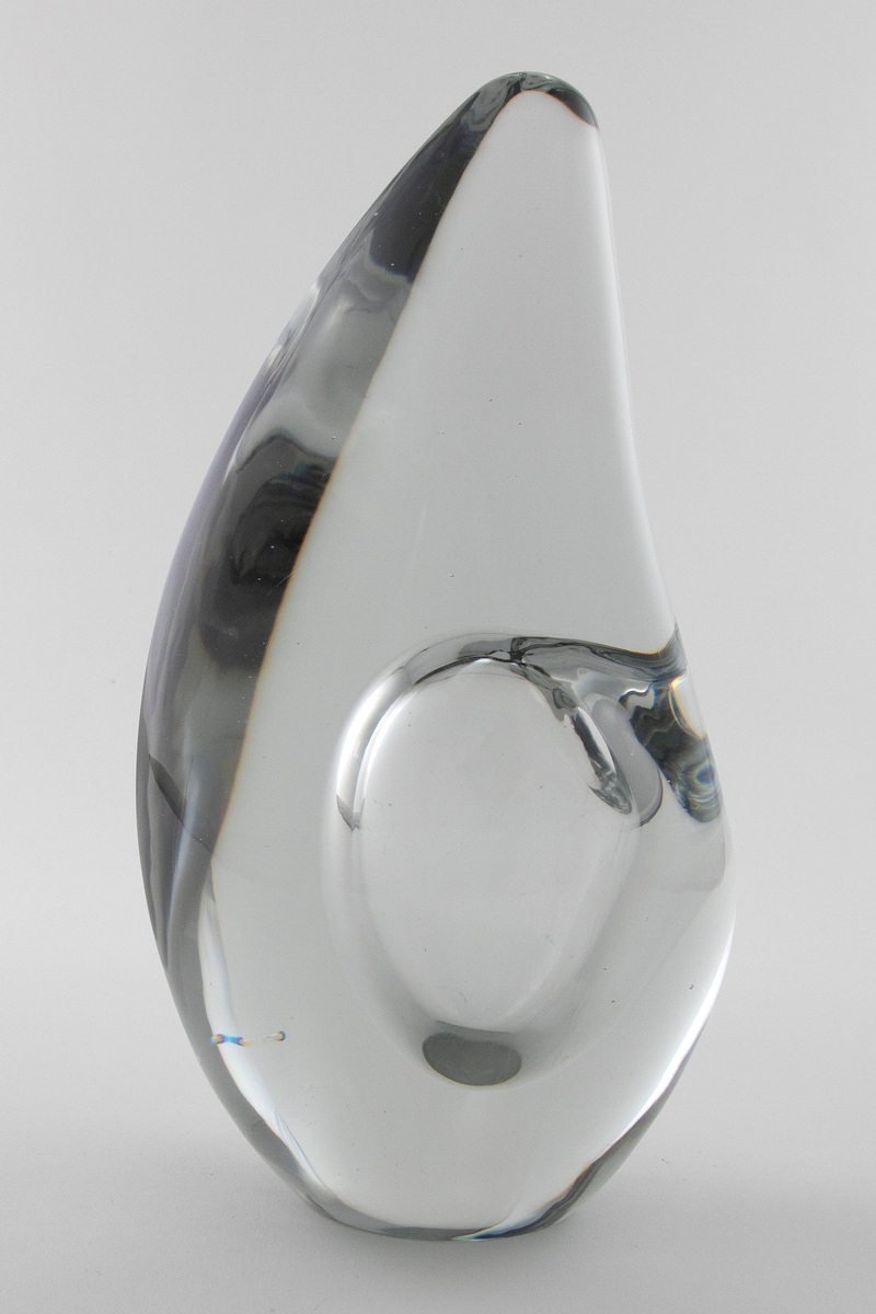 Dråpeformet skulpturell vase med abstrakt utforming i massivt klart glass. Et elipseformet hulerom i nedre del med liten sirkulær åpning. Bunnen er planslipt.