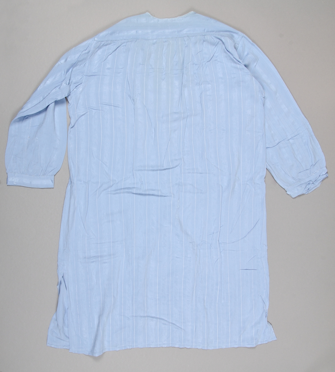 Nattskjorta av ljusblått blandat konstfibermaterial. Vävt med randig struktur med smala vita ränder. V-ringad utan krage. Knäppning framtill med två stora vita knappar.
Söndrig vid halslinningen. Nött.