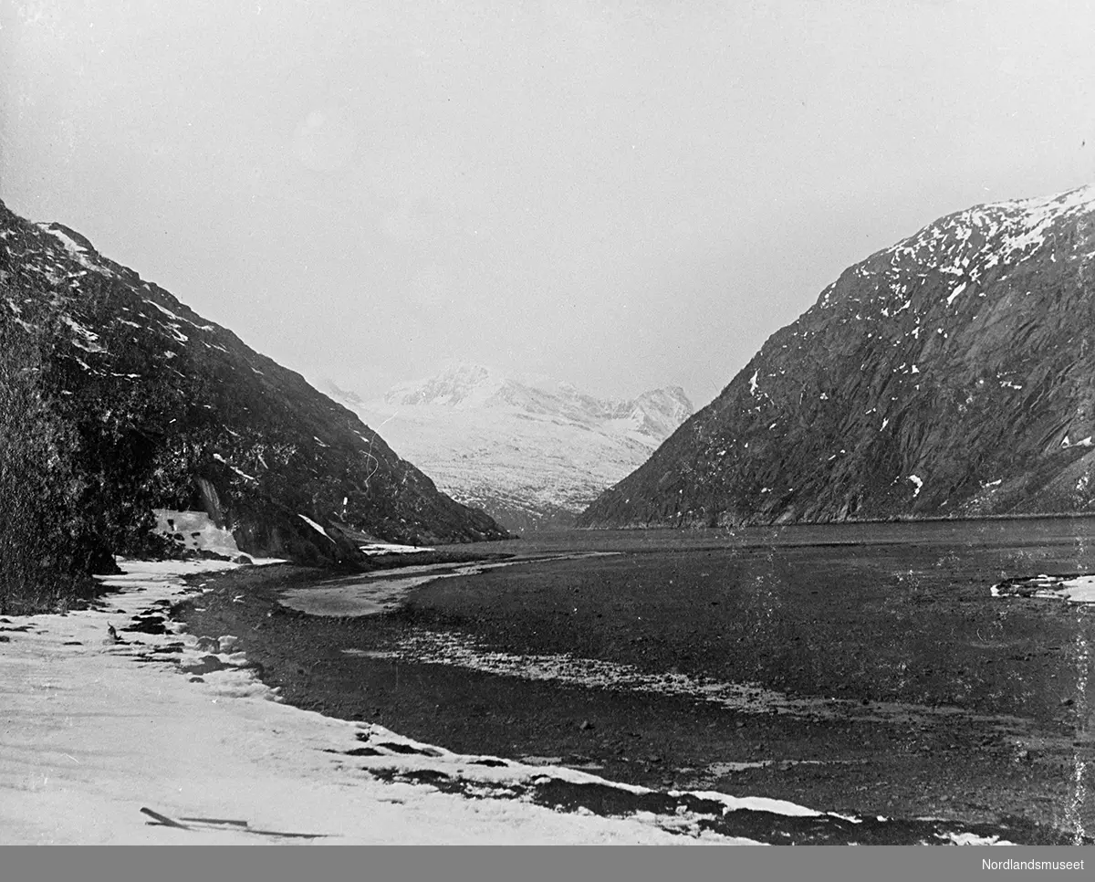 Narvik, Rombaksfjorden. Naturbilde. Strandkanten foran til venstre. Fjell på begge sider og bak. Liten odde til høyre. Noe av havet midt i bildet.